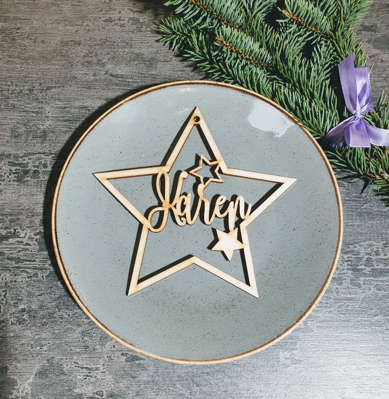 Puede incluir: Un adorno de madera en forma de estrella con el nombre "Karen" cortado en escritura cursiva. El adorno est&aacute; colocado sobre un plato gris con borde dorado, con una ramita de hoja perenne y una cinta morada en el fondo.
