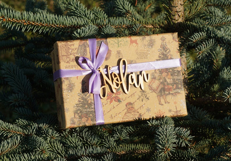 May include: A brown gift box with a purple ribbon and a wooden name tag that says "Nolan" on it. The box is sitting on a green pine tree branch.