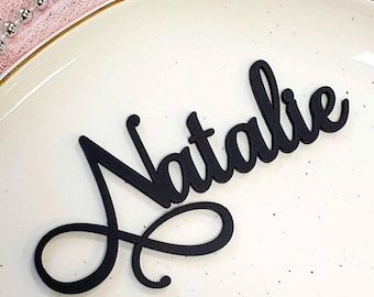 May include: A black, matte-finish name cutout in a cursive script font that spells the name Natalie. The lettering is positioned on a cream-colored plate with small black speckles and a thin gold rim, featuring a decorative silver beaded chain.