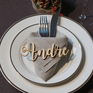 Puede incluir: Una tarjeta de lugar de madera con el nombre "Andr&eacute;" escrito en cursiva, colocada sobre una servilleta de lino doblada, con un tenedor y un cuchillo apoyados sobre la servilleta. La tarjeta de lugar est&aacute; sobre un plato blanco con un borde dorado.