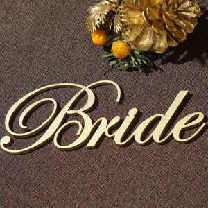 May include: A gold-colored wooden cutout of the word "Bride" in elegant script. The word is placed on a brown textured surface, with a decorative gold floral arrangement in the background. A wedding-themed item.