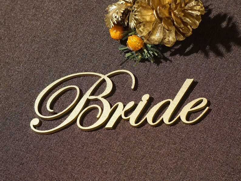 May include: A gold-colored wooden cutout of the word "Bride" in elegant script. The word is placed on a brown textured surface, with a decorative gold floral arrangement in the background. A wedding-themed item.
