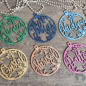 May include: Six round wooden ornaments with snowflake designs and personalized names. The ornaments are in various colors, including green, blue, pink, gold, and copper.