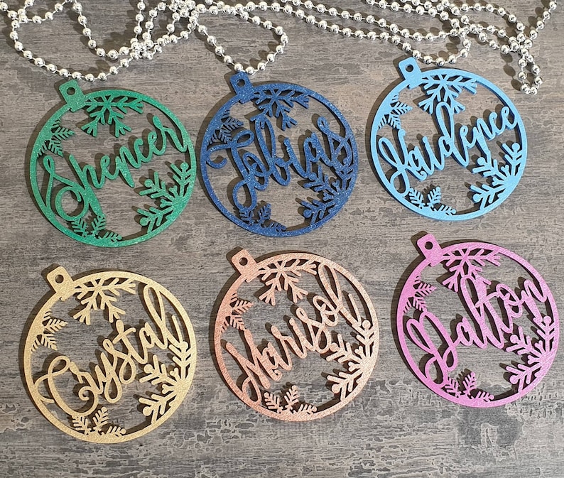 May include: Six round wooden ornaments with snowflake designs and personalized names. The ornaments are in various colors, including green, blue, pink, gold, and copper.