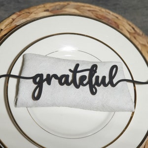 Pode incluir: Uma pe&ccedil;a de arte de palavras preta "grateful" em it&aacute;lico repousa sobre um guardanapo branco, colocado em um prato branco com borda dourada. O prato fica em cima de um tapete de mesa tecido de cor natural. Elemento decorativo para uma mesa.