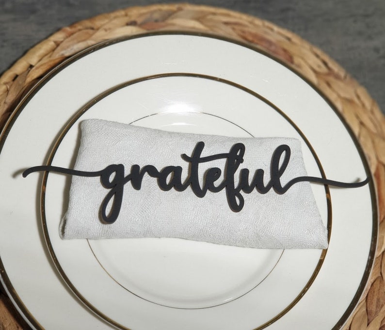 Pode incluir: Uma pe&ccedil;a de arte de palavras preta "grateful" em it&aacute;lico repousa sobre um guardanapo branco, colocado em um prato branco com borda dourada. O prato fica em cima de um tapete de mesa tecido de cor natural. Elemento decorativo para uma mesa.