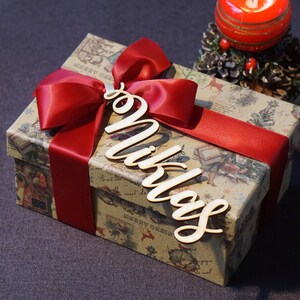 Puede incluir: Una caja de regalo marr&oacute;n con una cinta roja y una etiqueta de madera con el nombre "Niklas". La caja est&aacute; envuelta en papel con un tema navide&ntilde;o.