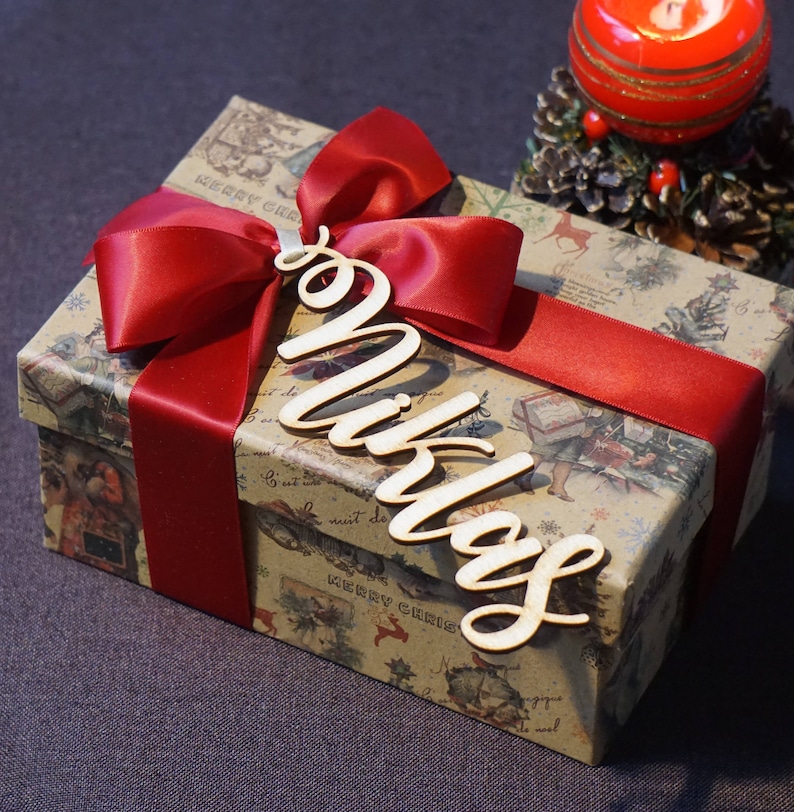 Puede incluir: Una caja de regalo marr&oacute;n con una cinta roja y una etiqueta de madera con el nombre "Niklas". La caja est&aacute; envuelta en papel con un tema navide&ntilde;o.