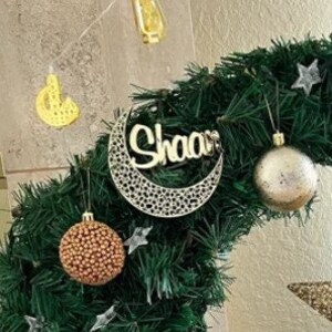 May include: A silver crescent moon ornament with the word "Shaan" written on it. The ornament is hanging on a green artificial Christmas tree with gold and silver ornaments.