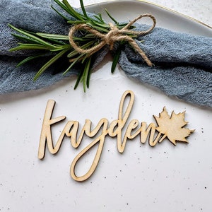 May include: Wooden place card with the name "Kayden" written in cursive script. A small wooden maple leaf is attached to the right side of the name.