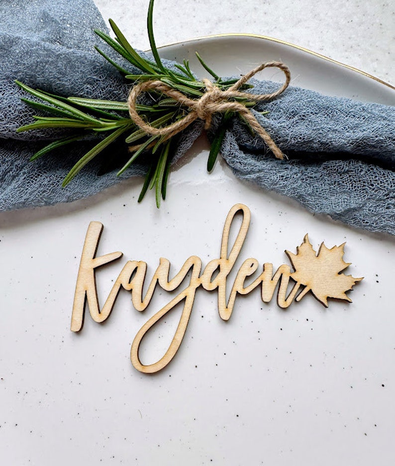 May include: Wooden place card with the name "Kayden" written in cursive script. A small wooden maple leaf is attached to the right side of the name.