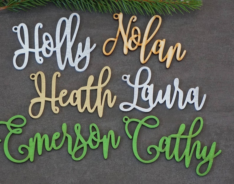 May include: Six wooden ornaments with names written in cursive. The names are Holly, Nolan, Heath, Laura, Emerson, and Cathy. The ornaments are painted in different colors: silver, gold, and green.