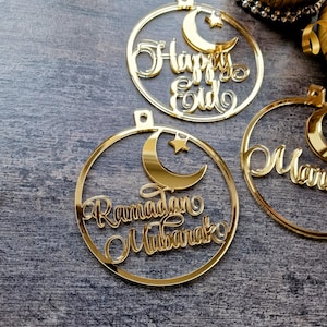 Op de afbeelding: Drie gouden, ronde, acryl ornamenten met een halve maan en ster ontwerp. De ornamenten hebben de tekst "Happy Eid", "Ramadan Mubarak" en "Mariam" erop geschreven.