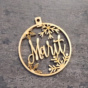 May include: A wooden Christmas bauble with the name "Marit" cut out in a cursive font. The bauble is circular with snowflake and pine branch cutouts around the edges. It has a small hole at the top for hanging.