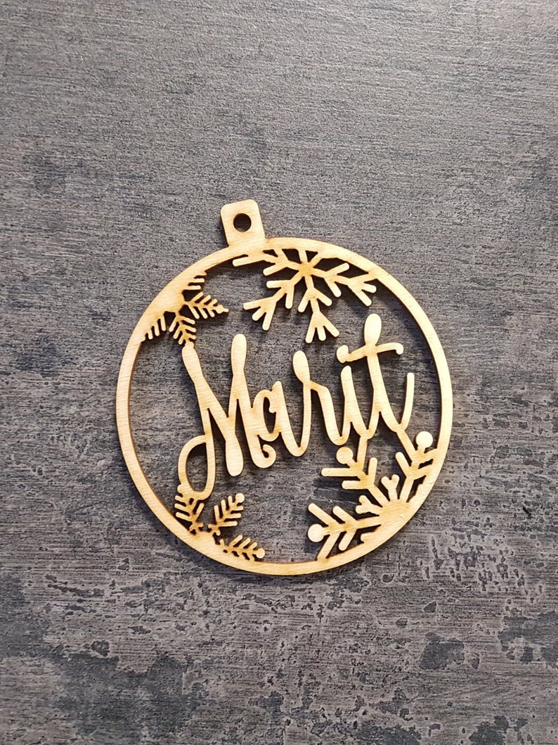 May include: A wooden Christmas bauble with the name "Marit" cut out in a cursive font. The bauble is circular with snowflake and pine branch cutouts around the edges. It has a small hole at the top for hanging.