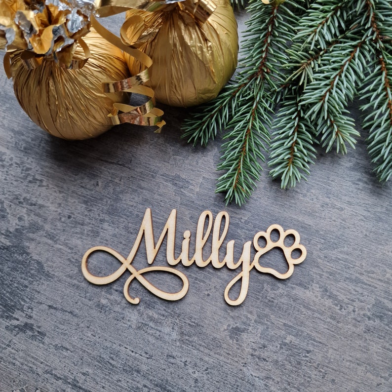 May include: A wooden sign with the name "Milly" and a paw print, surrounded by gold-wrapped gifts and a green pine branch.