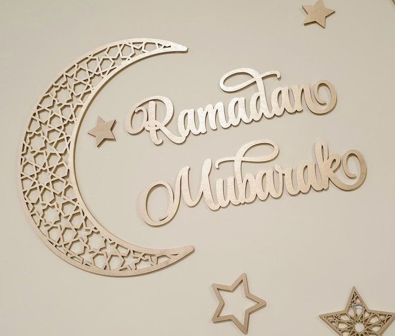 May include: A wooden cutout of a crescent moon with an intricate geometric pattern, along with the words "Ramadan Mubarak" in a decorative script. There are also several small wooden stars.