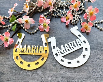 May include: Two gold and white horseshoe-shaped ornaments with the name "Maria" written on them. The ornaments are surrounded by pink flowers and a silver beaded garland.