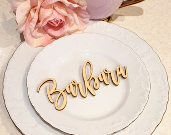 Puede incluir: Un plato de porcelana blanca con un borde dorado y una tarjeta de nombre de madera con el nombre "Barbara" escrito en cursiva.