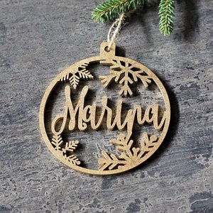 May include: A wooden Christmas ornament in the shape of a circle with a cutout design of snowflakes and the name "Marina" in a cursive font.