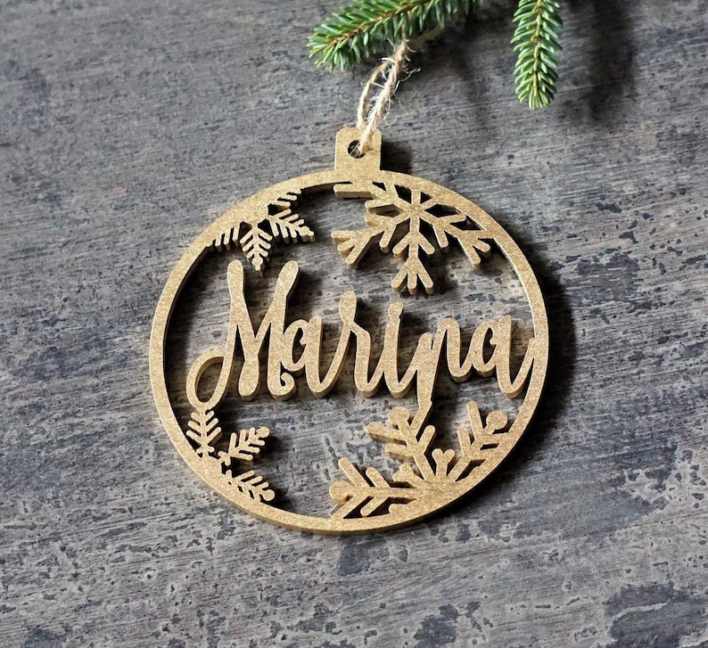 May include: A wooden Christmas ornament in the shape of a circle with a cutout design of snowflakes and the name "Marina" in a cursive font.