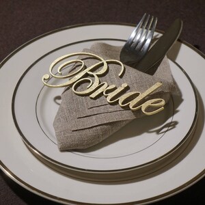 May include: A formal place setting featuring a white plate with a gold rim, a linen napkin, and gold cutlery. A gold-colored cursive "Bride" place card rests on the napkin, suggesting a wedding or bridal event. The overall aesthetic is elegant and celebratory.