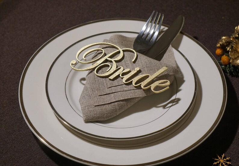 May include: A formal place setting featuring a white plate with a gold rim, a linen napkin, and gold cutlery. A gold-colored cursive "Bride" place card rests on the napkin, suggesting a wedding or bridal event. The overall aesthetic is elegant and celebratory.