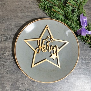 Puede incluir: Un adorno de madera en forma de estrella con el nombre "Karen" cortado en una fuente cursiva. El adorno tiene un dise&ntilde;o de estrella m&aacute;s peque&ntilde;o y un agujero para colgar. El adorno est&aacute; sobre un plato gris con un borde marr&oacute;n, con una ramita de hoja perenne y una cinta morada en el fondo.