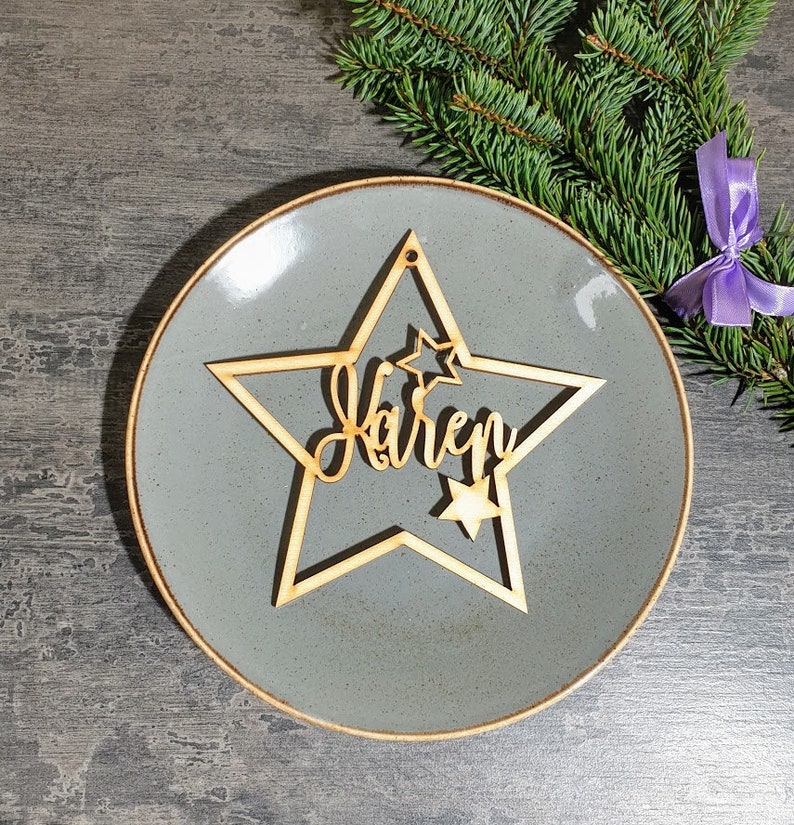 Puede incluir: Un adorno de madera en forma de estrella con el nombre "Karen" cortado en una fuente cursiva. El adorno tiene un dise&ntilde;o de estrella m&aacute;s peque&ntilde;o y un agujero para colgar. El adorno est&aacute; sobre un plato gris con un borde marr&oacute;n, con una ramita de hoja perenne y una cinta morada en el fondo.