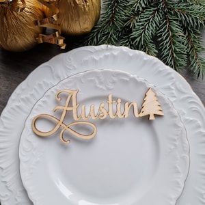 May include: A wooden place card with the name "Austin" written in cursive script. A small wooden Christmas tree is attached to the right side of the card. The place card is sitting on a white plate.