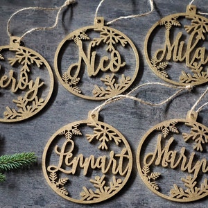 May include: Five wooden Christmas ornaments with names Leroy, Nico, Mika, Lennart, and Maryna written in cursive on each ornament. The ornaments are decorated with snowflakes.
