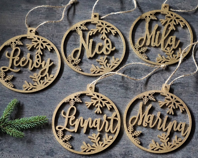 May include: Five wooden Christmas ornaments with names Leroy, Nico, Mika, Lennart, and Maryna written in cursive on each ornament. The ornaments are decorated with snowflakes.