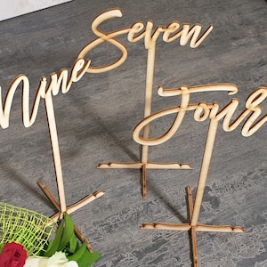 May include: Three wooden table number signs. The numbers are in a cursive font and read "Nine", "Seven", and "Four". Each number is attached to a stand with a cross-shaped base. The signs are made of light-colored wood.