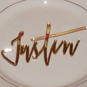 May include: Gold mirror acrylic name card with the name "Justin" written in a cursive script.