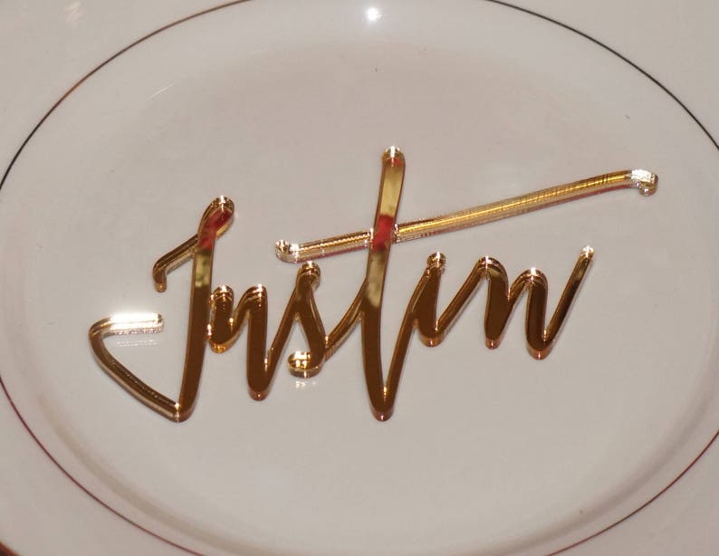 May include: Gold mirror acrylic name card with the name "Justin" written in a cursive script.