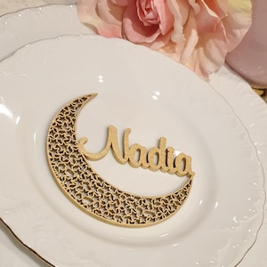 May include: A wooden crescent moon with a star pattern and the name Nadia written on it. The moon is on a white plate with a gold rim.