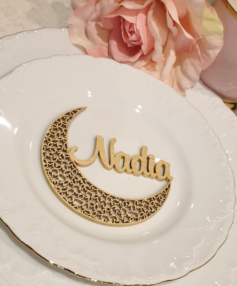 May include: A wooden crescent moon with a star pattern and the name Nadia written on it. The moon is on a white plate with a gold rim.