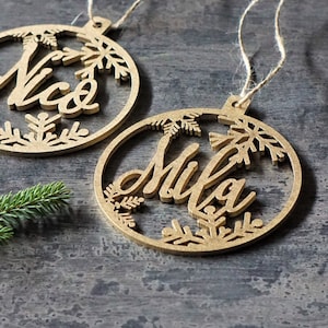 May include: Two wooden Christmas ornaments with the names "Nico" and "Mila" cut out in the center. The ornaments are decorated with snowflake designs.