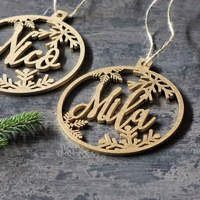 May include: Two wooden Christmas ornaments with the names "Nico" and "Mila" cut out in the center. The ornaments are decorated with snowflake designs.