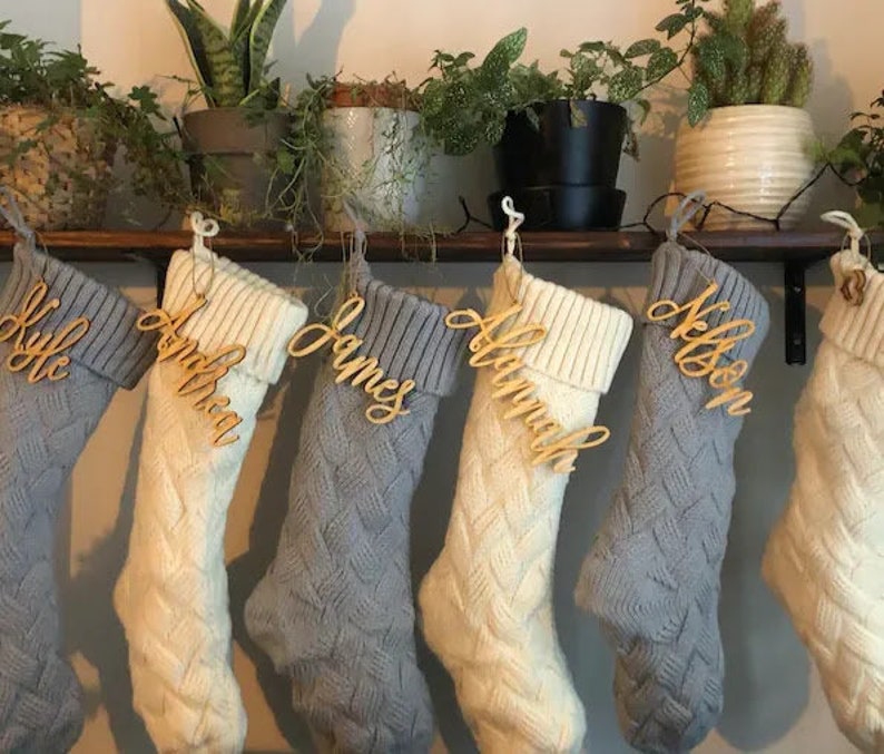 May include: Six white and gray knitted stockings with wooden name tags. The names on the tags are Kyle, Amber, James, Amanda, Nelson, and a sixth name that is not visible.