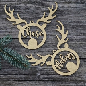 May include: Two wooden reindeer head ornaments with the names "Chase" and "Mabry" cut out. The ornaments are light brown and have antlers. A sprig of blue-green pine needles is in the corner. The ornaments are on a gray wooden surface.