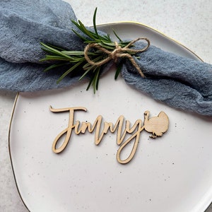 May include: A wooden Thanksgiving place card with the name "Jimmy" written in cursive and a turkey silhouette. The place card is on a white plate with a blue napkin and a sprig of rosemary.