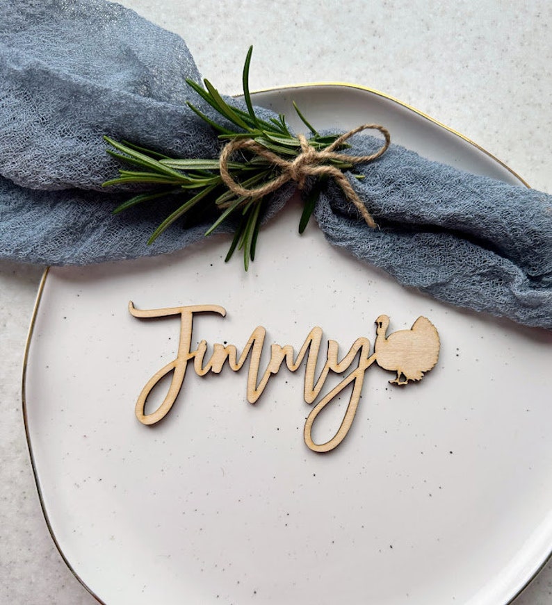 May include: A wooden Thanksgiving place card with the name "Jimmy" written in cursive and a turkey silhouette. The place card is on a white plate with a blue napkin and a sprig of rosemary.