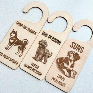 May include: Three light-colored wooden door hangers with hook tops. Each features a laser-engraved dog illustration and text. The first reads HUND IM ZIMMER TE NICHT STÖREN, the second DOG IN ROOM! PLEASE DO NOT DISTURB!, and the third SUNS LŪDZU PIEKLAUVĒT.