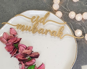 Può includere: Un piatto bianco con un bordo dorato, decorato con un cartello di legno che recita "eid mubarak". Il piatto è circondato da petali di fiori rosa e una ghirlanda di luci bianche.
