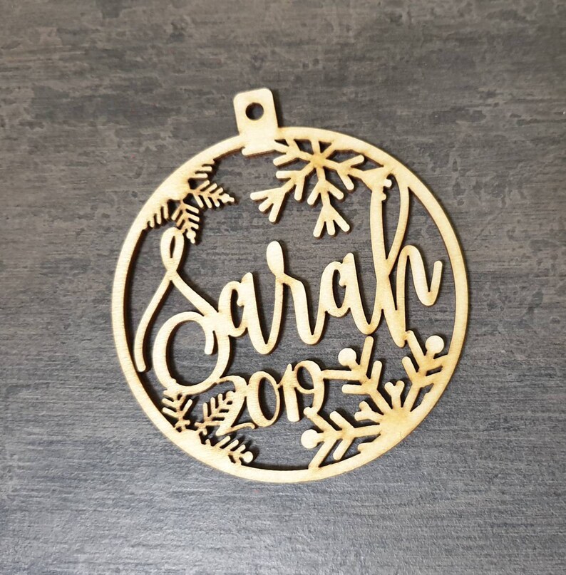 May include: A wooden Christmas ornament with a snowflake design and the name "Sarah" and the year "2019" cut out.
