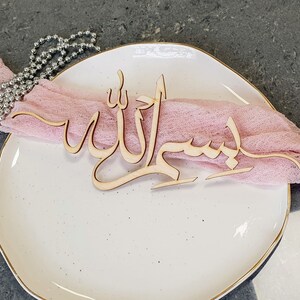 Op de afbeelding: Een houten uitsnijding van de Arabische zin "Bismillah" (In de naam van Allah) op een wit bord met een roze stoffen servet.