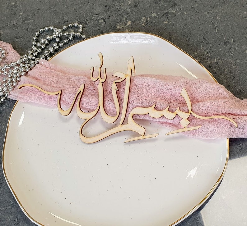Op de afbeelding: Een houten uitsnijding van de Arabische zin "Bismillah" (In de naam van Allah) op een wit bord met een roze stoffen servet.