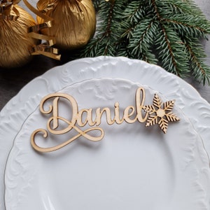 May include: Wooden name card with the name "Daniel" written in cursive script. A wooden snowflake is attached to the right side of the name. The name card is placed on a white plate.