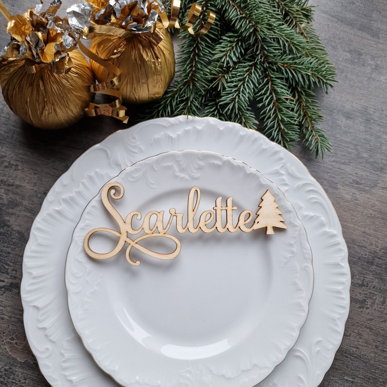 May include: A white plate with a wooden name card that says "Scarlette" and a small wooden Christmas tree. The plate is on a wooden table with two gold wrapped gifts.